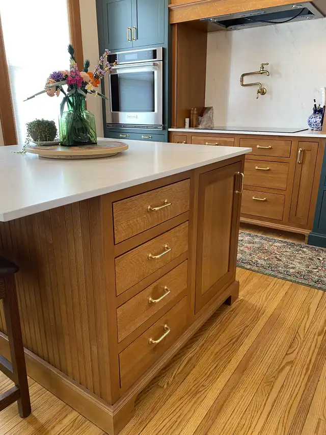 Kitchen island with stained wood cabinetry and a white quartz top in front of deep green cabinets and a brass pot filler