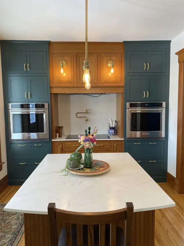 Deep green painted cabinets around double wall ovens with a stained wood hutch, brass hardware and a quartz island